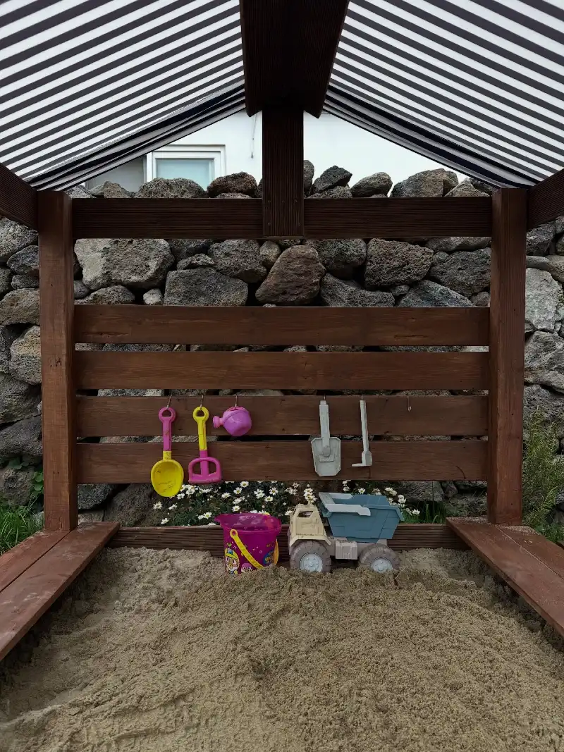 Dark-Stained Wooden Sandbox with Striped Canopy Against a Rustic Stone Wall